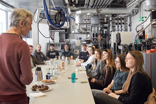 Eine Gruppe Studierender sitzt an einem großen Tisch, auf dem Snacks und Getränke stehen. Im Vordergrund ist ein Dozent zu sehen, der zu ihnen spricht.